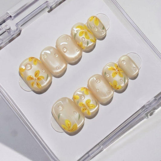 EyeCandys After Rain Press-On Nails feature a pearly base with yellow flower designs and white dots, presented in a clear case—ideal for achieving a salon-quality gel manicure at home with reusable nails.