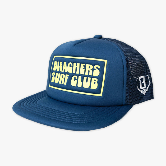 A navy blue trucker hat with a yellow outlined 'BLEACHERS SURF CLUB' patch on the front, featuring a navy mesh back and a small white logo on the side.