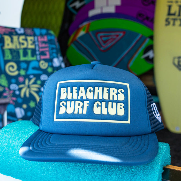 A blue trucker hat with 'BLEACHERS SURF CLUB' printed in white on the front, displayed on a turquoise stand with surfboards in the background.