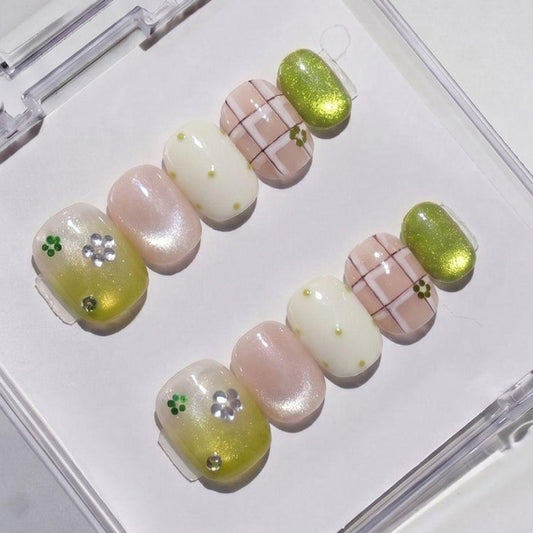 The EyeCandys Garden Variety Press-On Nails set features handmade designs in green, white, and nude with plaid, metallic green, flowers, rhinestones, and dots—neatly arranged in a plastic case for a salon gel look.
