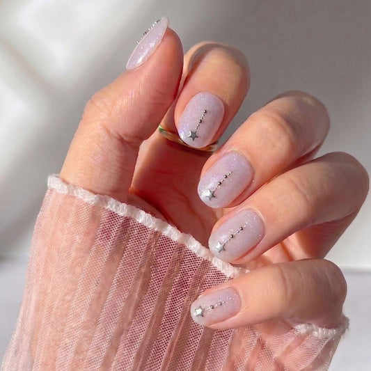 A hand wears EyeCandys Gemini Press-On Nails—light pink, sheer, and glossy with tiny silver star and dot accents. The wrist and fingers are draped in sheer, textured light pink fabric.