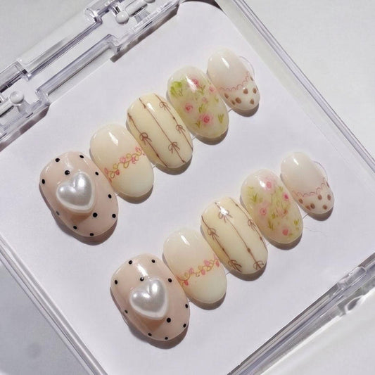 Little Bea Press-On Nails by EyeCandys feature pastel cottagecore designs—bows, tiny flowers, dots, scalloped edges, and two nails with large 3D pearl hearts—all handmade and arranged in a clear case for a salon gel manicure look.