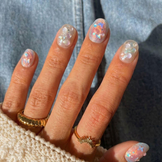 A hand shows off EyeCandys Love Tomorrow Press-On Nails—short, rounded with iridescent, holographic nail art, pearls, and star accents. Gold rings and a cream knit sweater pop against a denim background for a chic salon-style look.