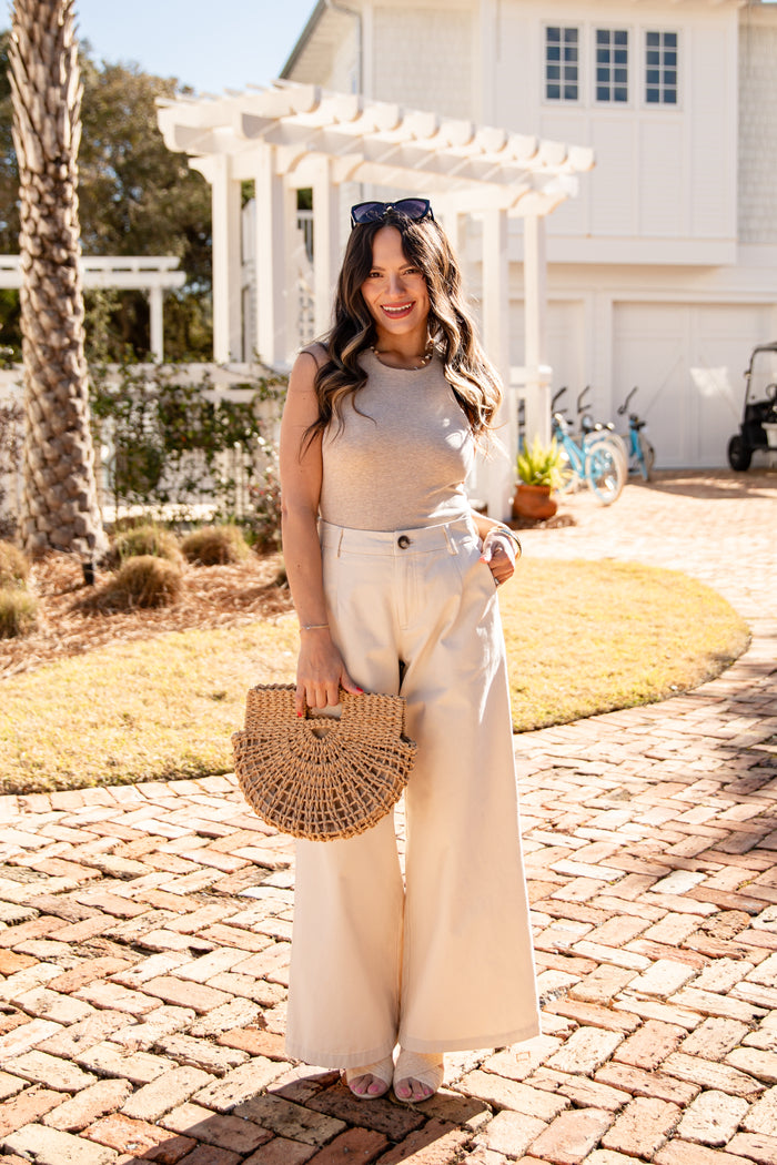 Woman standing on a brick path holding a woven bag, wearing a beige top and white pants, with a house and garden in the background.
