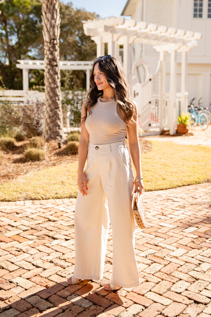 Woman in beige top and white pants standing on a brick path with a white building and trees in the background.