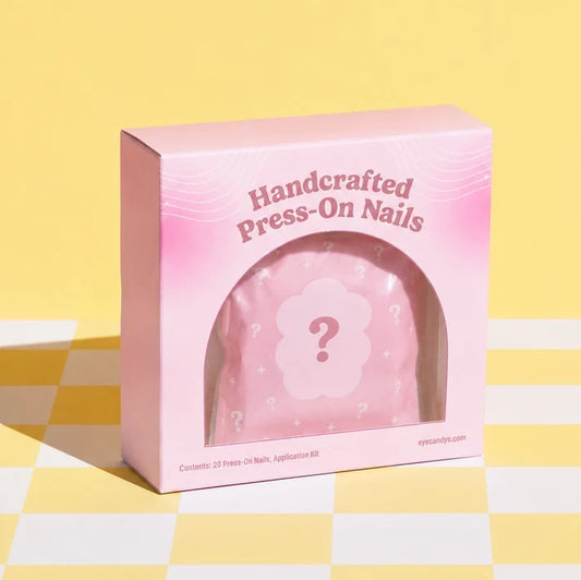 Handcrafted Press-O Nails mystery blind box in a pink cosmetic box with a frosted mystery pouch visible through an arched window on a pastel yellow checkered background.