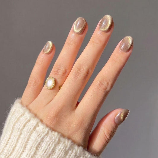 A hand wearing Velvet Taupe Press-On Nails by EyeCandys, shaped short and oval. The nails have holographic gold glitter. The person wears a gold ring with a white stone and a cream knit sweater against a plain gray background.